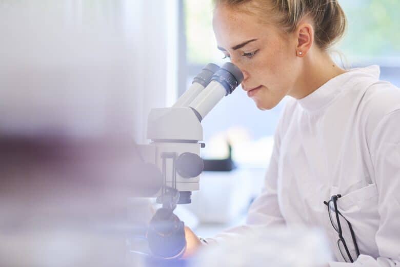 a female research scientist is analysing a sample on her microscope in a microbiology lab .  the lab is brightly lit with natural light . Blurred glassware at side of frame provides copy space .
